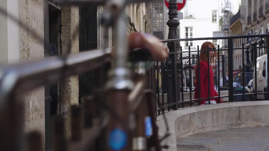 A stylish young woman with red hair walks out of a narrow alleyway onto a cobblestone street in Paris in slow motion