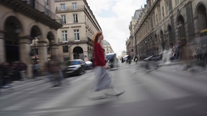 A woman in a red sweater stands out from the motion-blurred crowd as she crosses a busy street in Paris