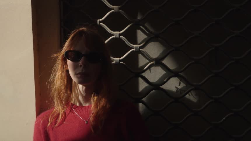 A stylish woman with red hair and sunglasses stands in a sliver of sunlight next to a dark metal gate