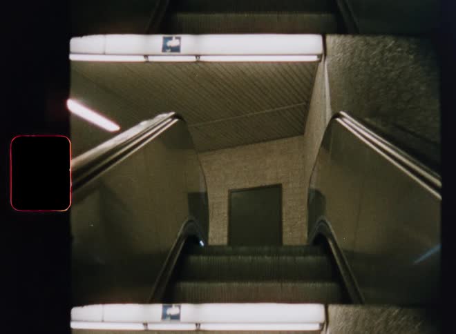 A surreal, kaleidoscopic view from a vintage camera descends an empty escalator into an underground subway station