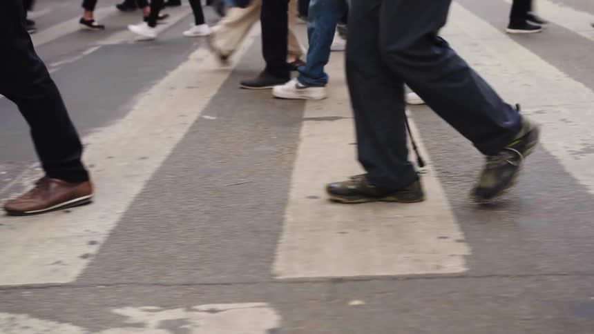A slow-motion, low-angle shot of a crowd's legs and feet as they walk across a city street crosswalk