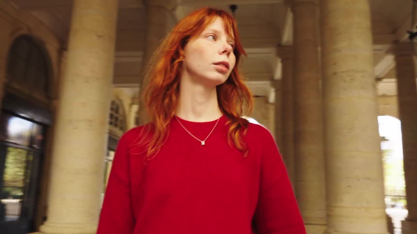 A beautiful young woman with red hair walks thoughtfully through a classical stone arcade in slow motion
