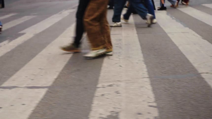 Low-angle panning shot of pedestrians' legs and feet walking across a city street crosswalk