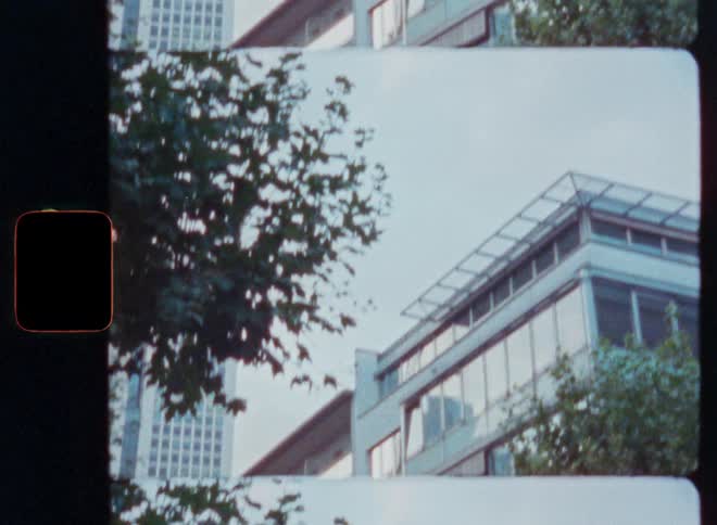 Vintage 8mm film scan reveals modern city buildings and skyscrapers framed by trees with a red light burn