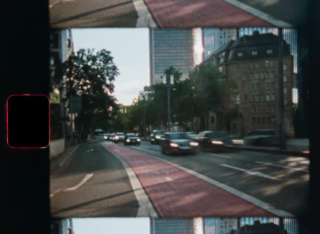A gritty 8mm film scan shows motion-blurred traffic on a bustling city street with a cyclist riding down a prominent red bike lane