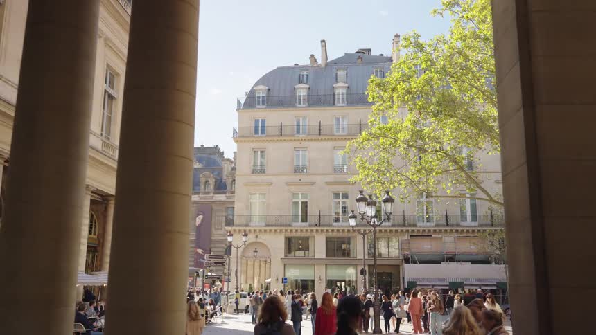 Slow reveal shot moving from a shadowed colonnade into a bright, bustling Parisian city square filled with pedestrians