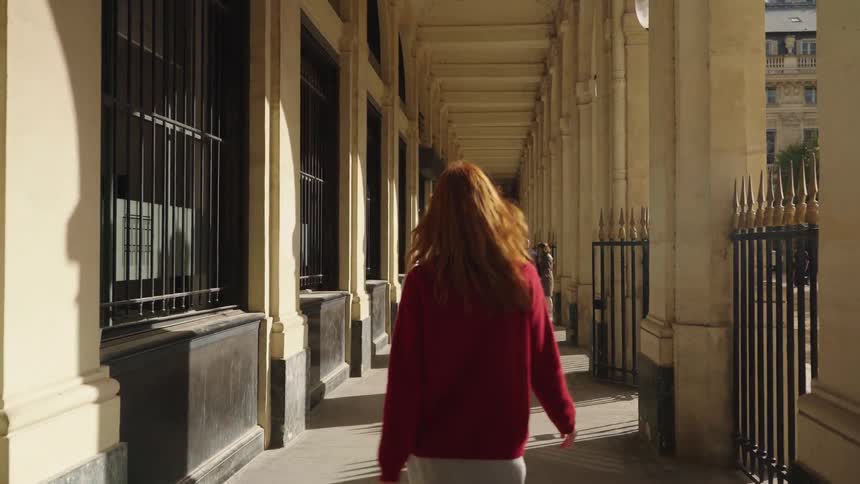 A chic woman with long red hair gracefully walks down a historic sunlit arcade in slow motion