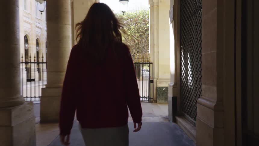 In slow motion, a stylish young woman with red hair walks down an elegant arched stone colonnade