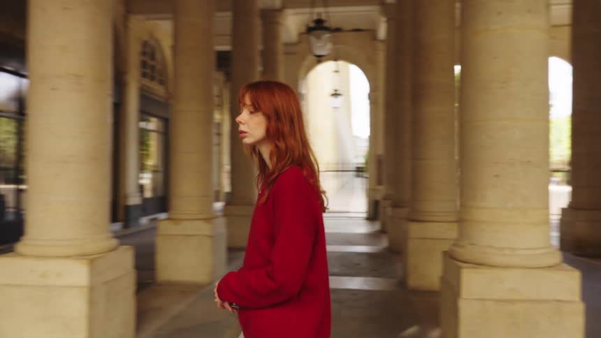 A pensive young woman with red hair walks in slow motion through a grand stone colonnade in a European city