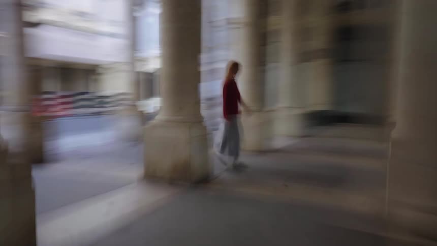 A woman wearing a red sweater walks through a stone colonnade in an artistically blurred and disorienting clip