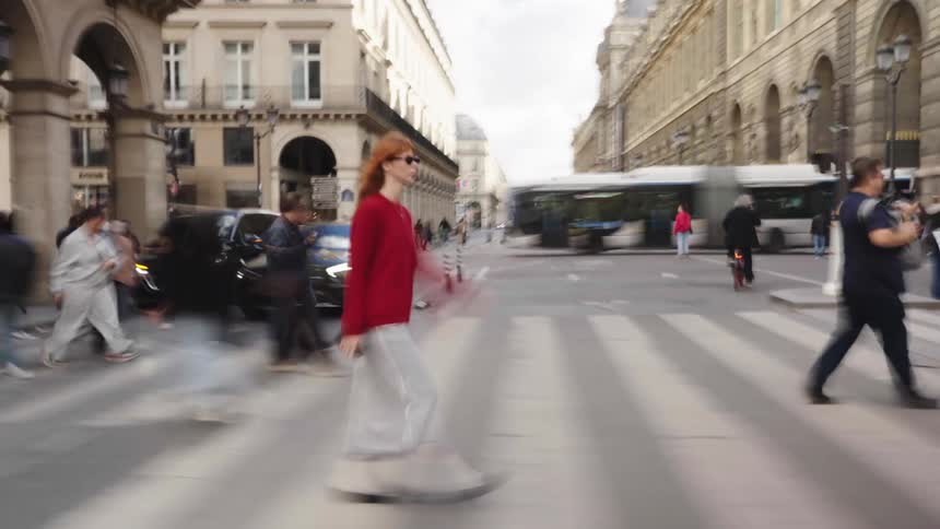 A woman in a red shirt walks in focus across a busy city street while the surrounding pedestrians and traffic move in a motion blur