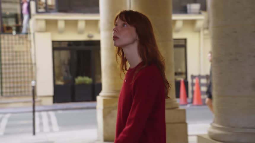 A contemplative young woman with flowing red hair walks in slow motion along a stone colonnade in a classic European city