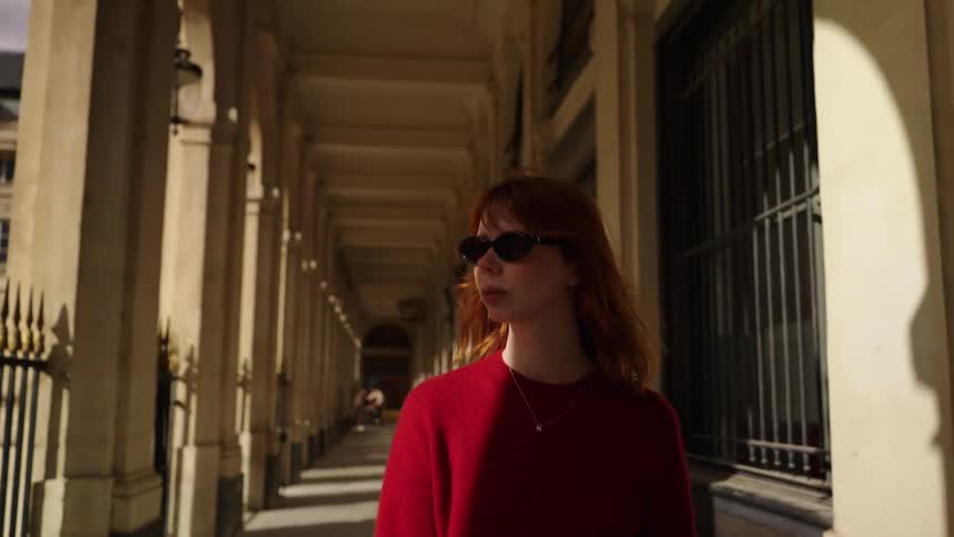 A stylish woman with red hair walks in slow motion through a sunlit arcade with stone columns