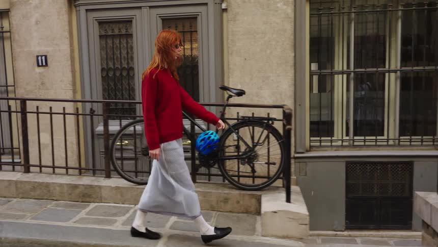 A stylish redhead in a red sweater and long skirt walks in slow motion along a picturesque European street