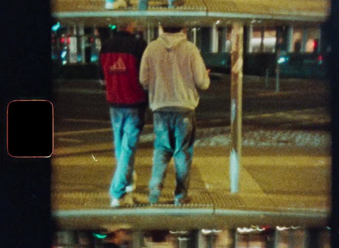 A vintage, reflective film shot captures two young men from behind walking on a city street at night