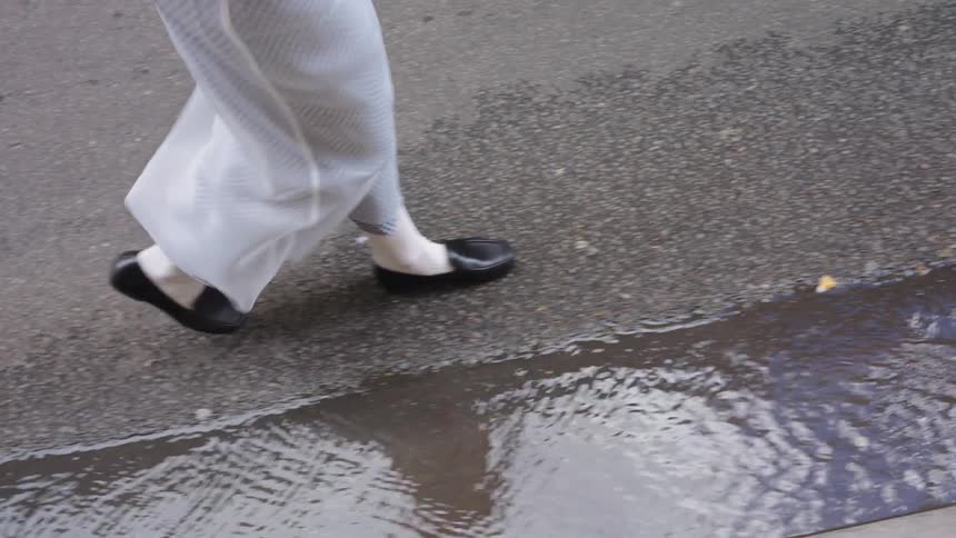 Slow motion close-up of a person's feet in traditional attire walking on a wet sidewalk next to a puddle
