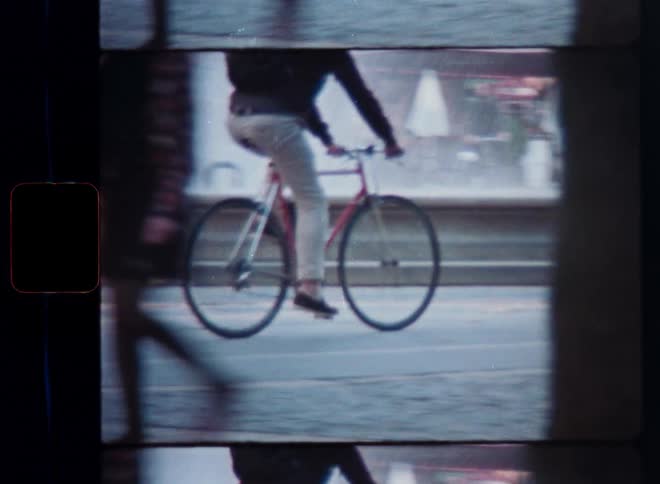 A vintage film shot tracks a cyclist riding a red bicycle through a bustling city square