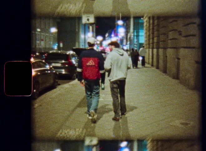 Handheld vintage film shot of two young men walking down a city street at night, with streetlights and cars in the background