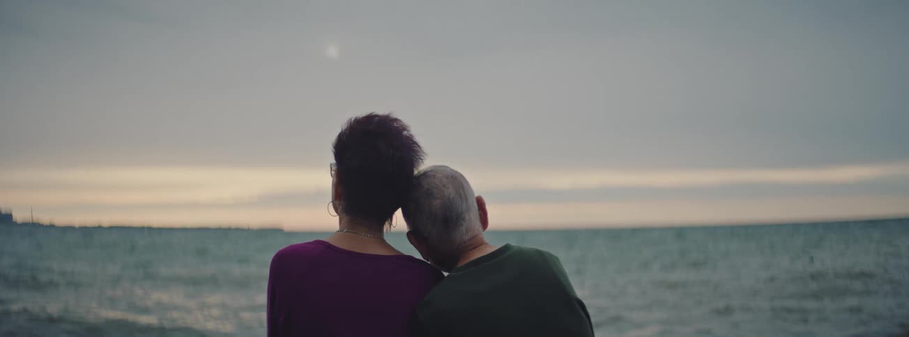 Back view of a loving senior couple sitting together by the water as the man rests his head on the woman's shoulder