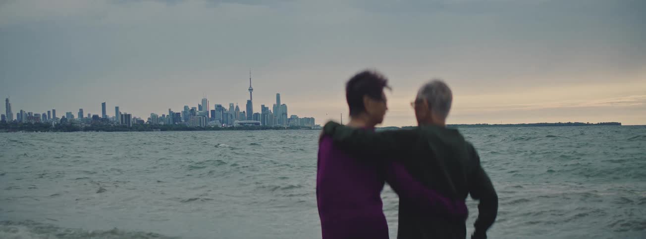 A senior couple with their arms around each other stand on a beach looking out at the Toronto skyline across the water