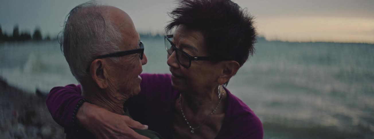 An intimate close-up shows a senior Asian couple embracing and looking into each other's eyes on a beach at twilight
