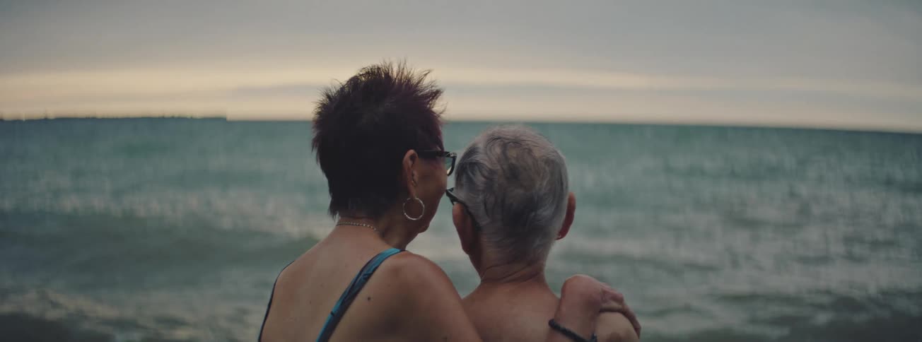 A loving senior couple stands together with their backs to the camera, looking out at the calm ocean at dusk
