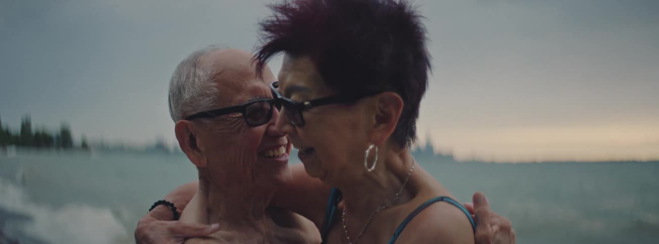 A loving elderly couple in swimsuits smiles and laughs while sharing a tender embrace by the ocean