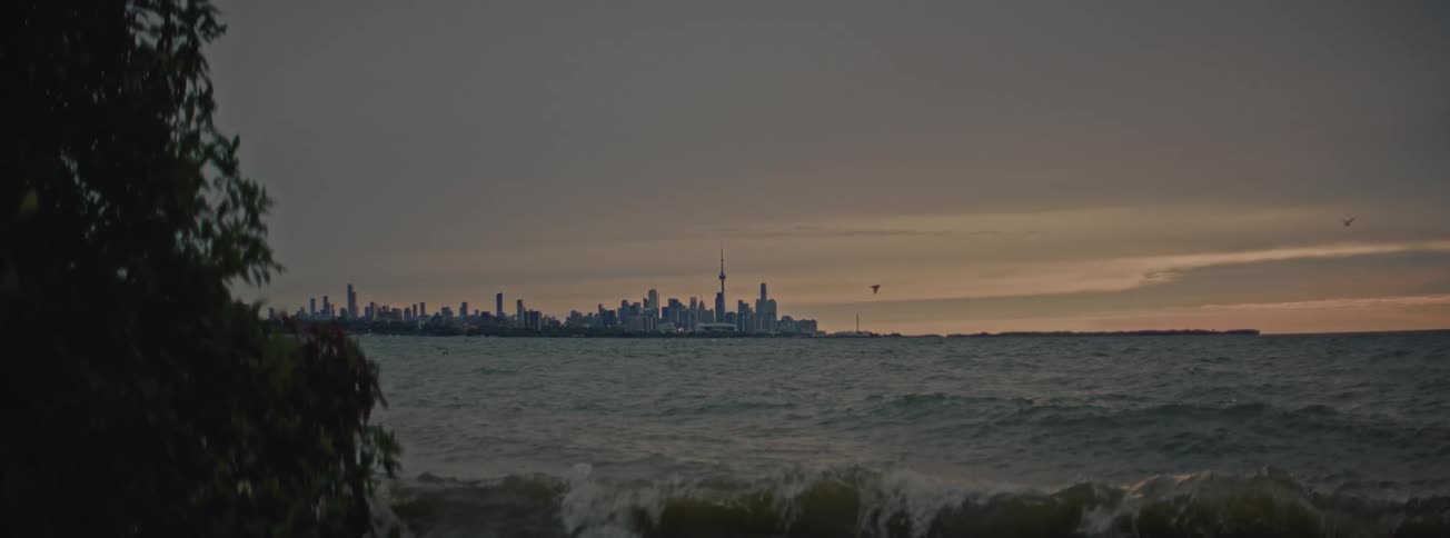A distant view of the Toronto skyline over choppy waters, with waves breaking on the shore in slow motion during a cloudy sunset