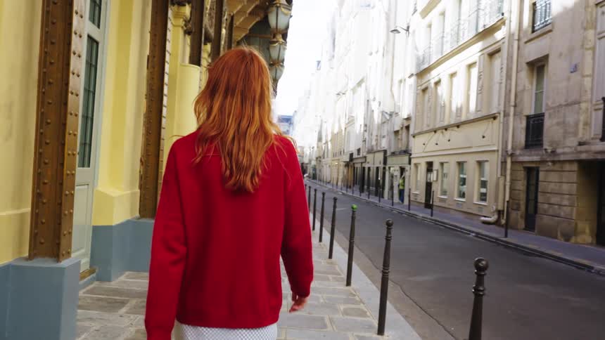 A stylish woman with red hair walks down a charming, narrow Parisian street in slow motion