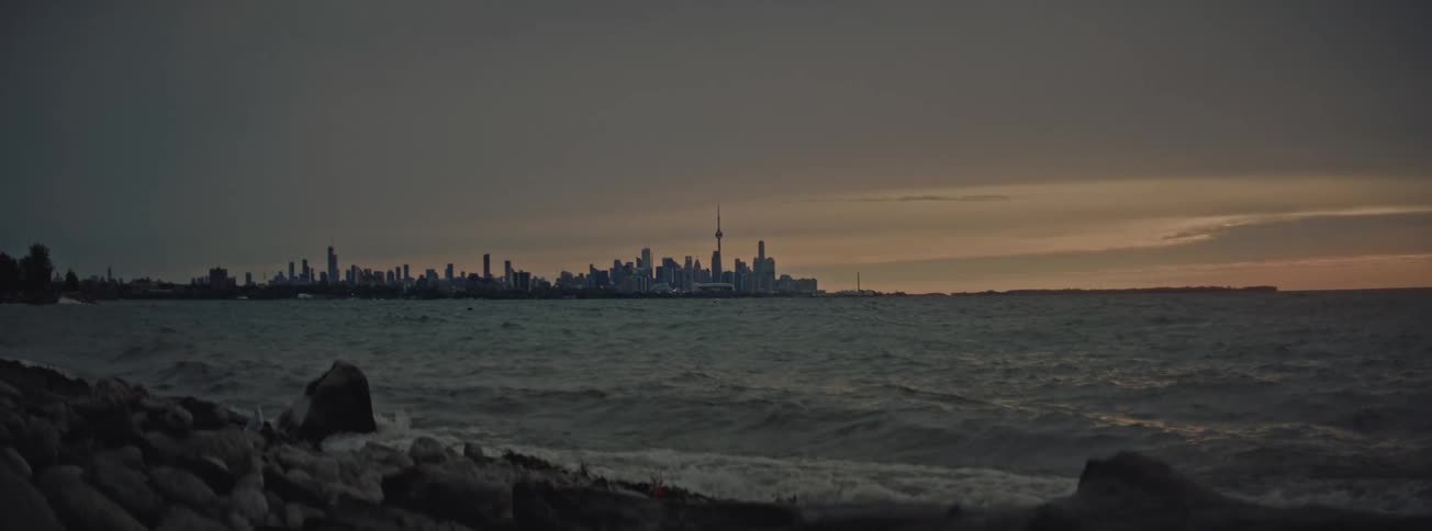 Slow motion waves crash on a rocky shore with the Toronto city skyline visible across the water under an overcast sky at dusk