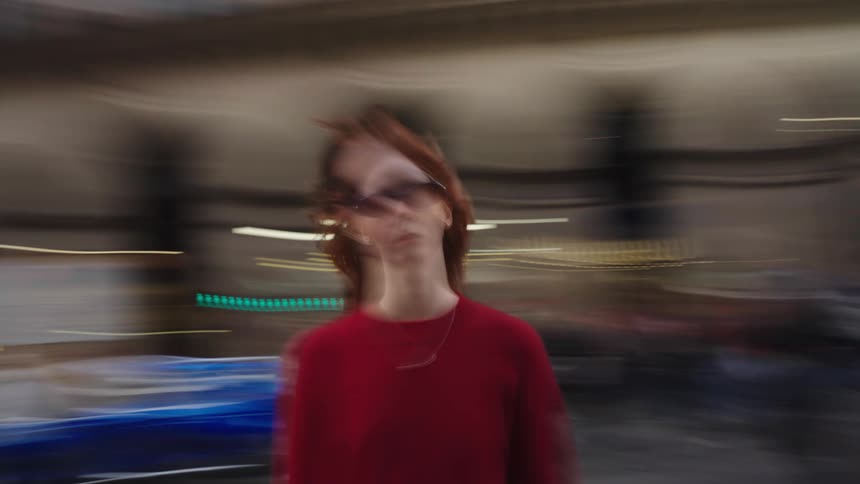 A woman with red hair stands still in the middle of a busy street as the city and crowds move around her in a motion-blurred time-lapse