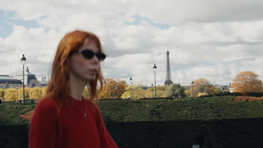 Slow motion shot of a stylish woman with red hair walking through the Tuileries Garden in Paris with the Eiffel Tower in the background