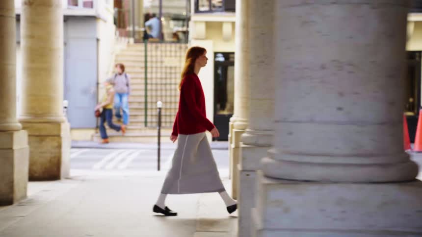 A stylish young woman with red hair walks in slow motion through a classic European colonnade in a seamless loop