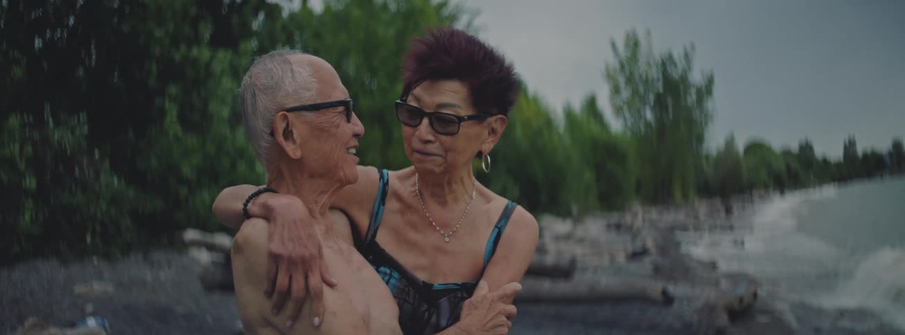 A loving senior couple in swimwear and sunglasses embrace and talk while sitting on a rocky beach