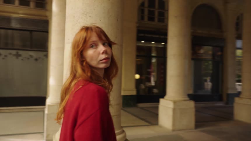 A stylish young woman with long red hair walks in slow motion through a classic European colonnade, looking back thoughtfully