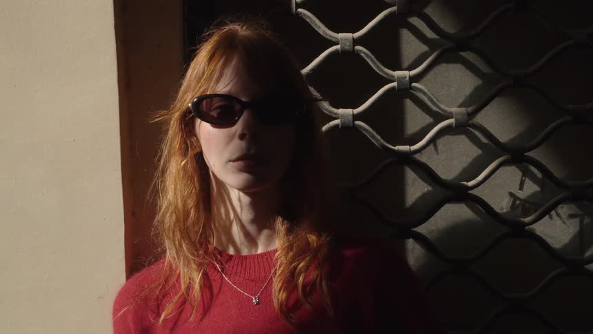 A stylish redhead in sunglasses and a red sweater slowly moves from the shadows into a ray of sunlight against a metal gate
