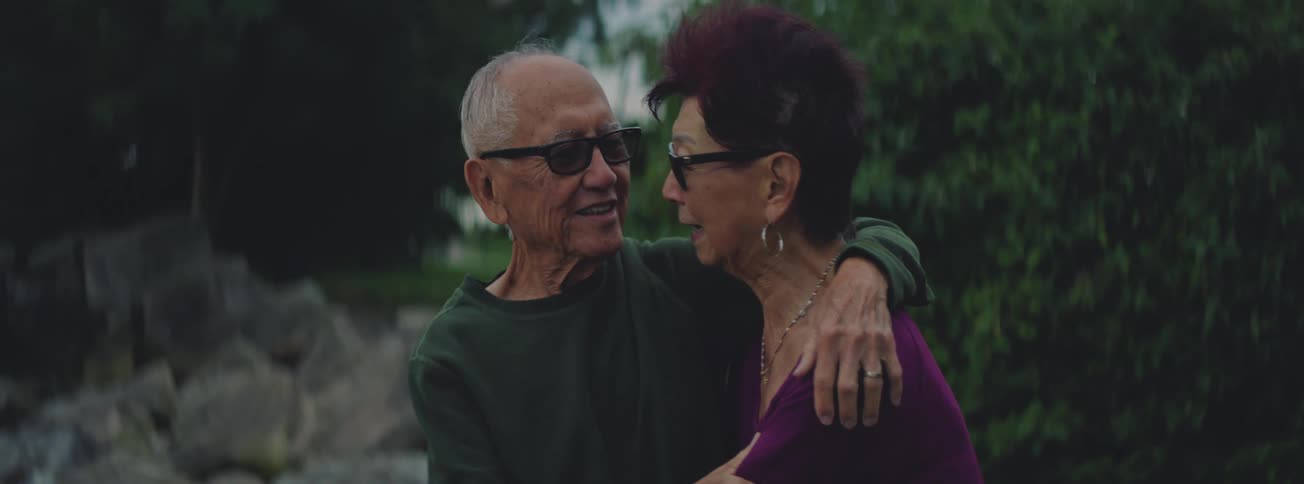 An elderly man and woman wearing sunglasses share a loving and happy conversation while embracing in a park