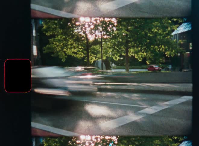 Authentic vintage film captures a time-lapse of cars blurring down a sunny street lined with lush green trees