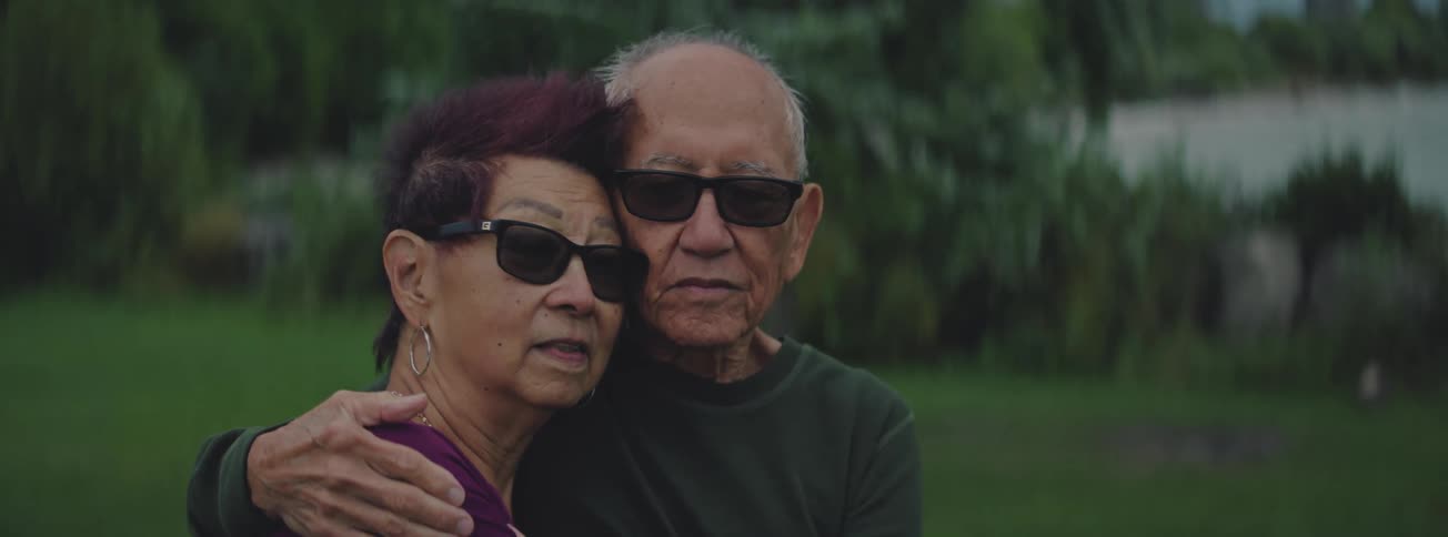 A stoic elderly couple wearing sunglasses embrace each other in a quiet outdoor portrait