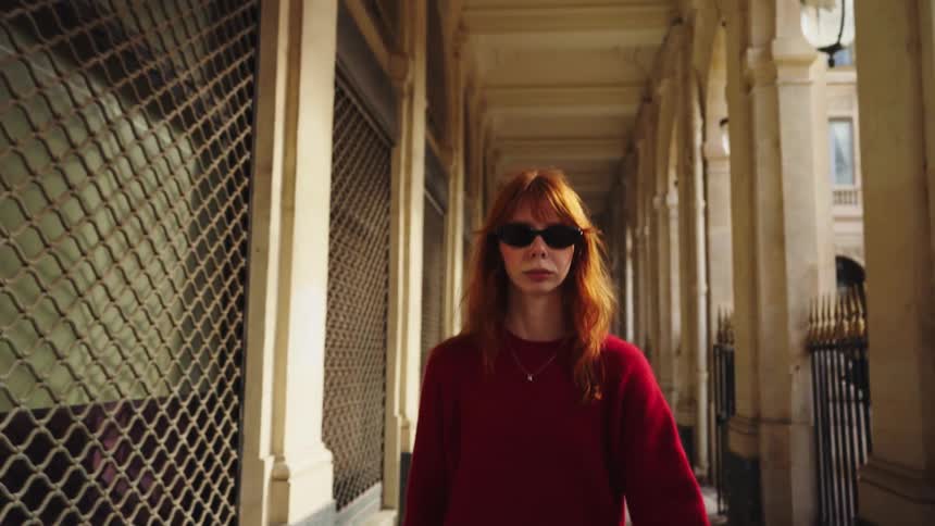 A stylish young woman with red hair and sunglasses walks confidently towards the camera in slow motion along a classic European colonnade