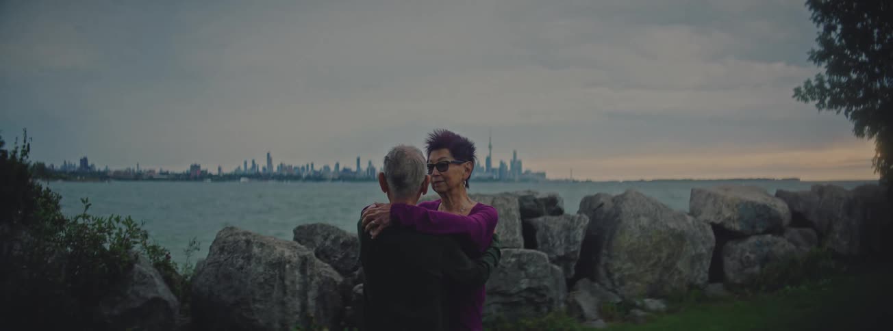 A loving senior couple embraces and sways together on the coast with a distant city skyline under an overcast sky