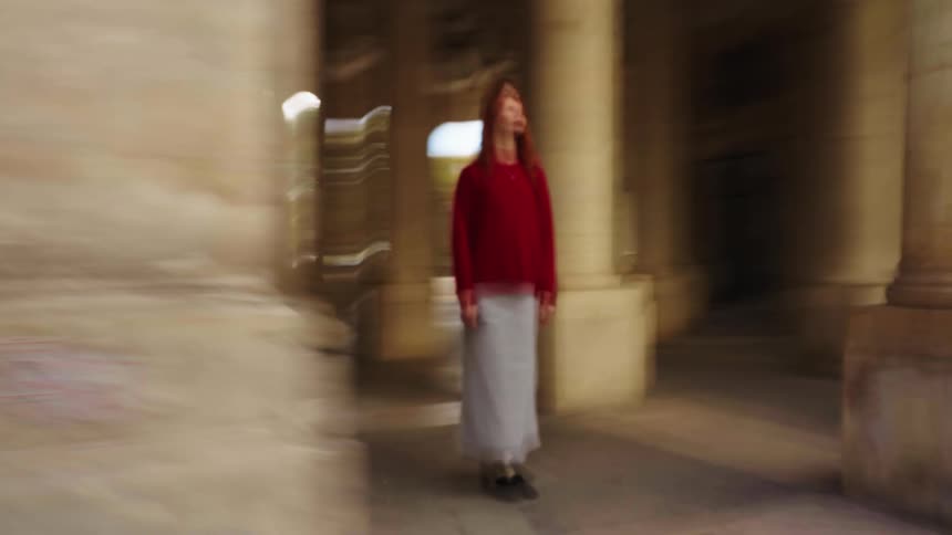 A woman in a red sweater stands still in a dreamy, motion-blurred colonnade as the camera pans quickly around her