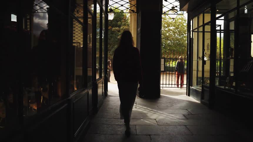 A silhouetted woman walks slowly away from the camera through a dark city passageway and into the bright sunlight