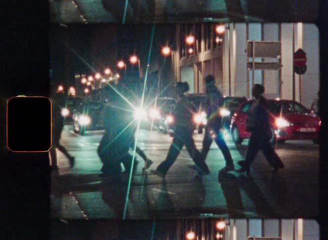 Gritty Super 8mm style footage captures silhouetted pedestrians crossing a busy city street at night as car headlights flare