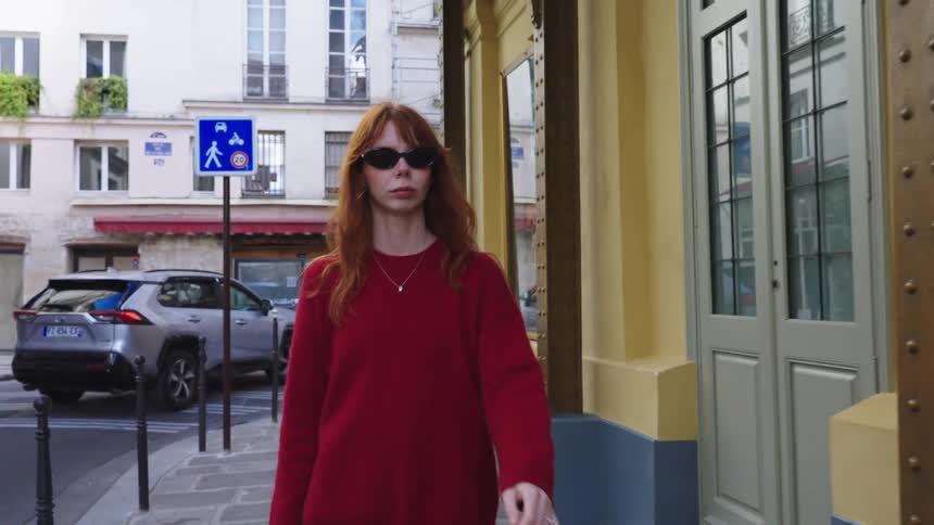A stylish young woman with red hair and sunglasses walks confidently down a Parisian street in slow motion
