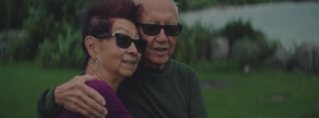 A slow motion close-up reveals an elderly Asian couple wearing sunglasses sharing a tender embrace outdoors
