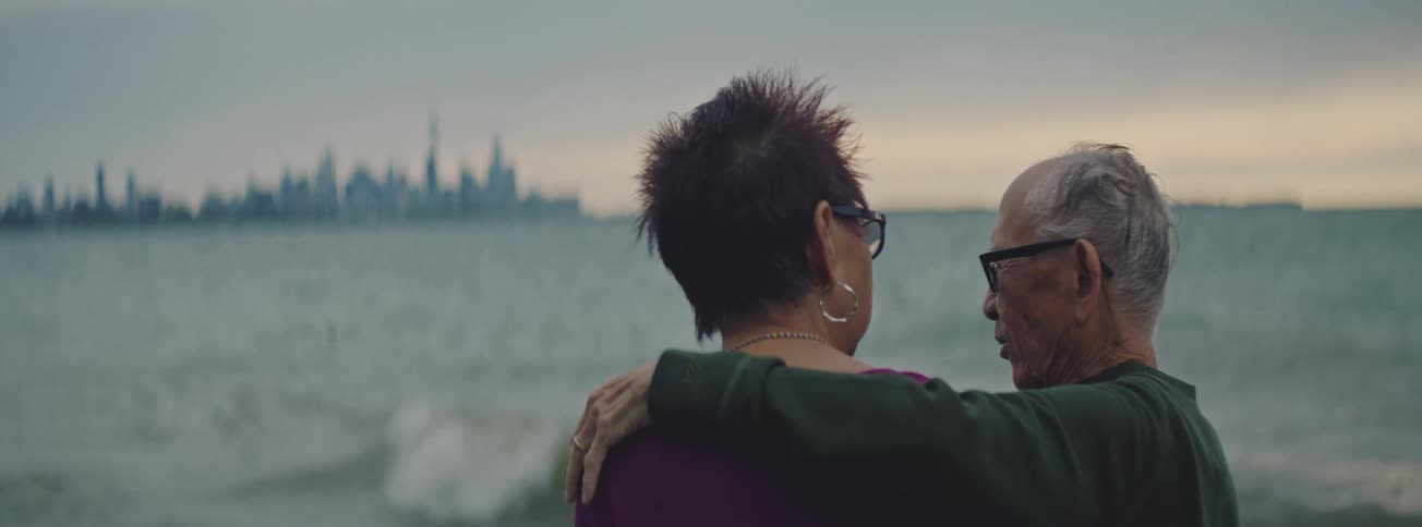 An elderly couple embraces while watching the waves roll in, with a distant city skyline in the background