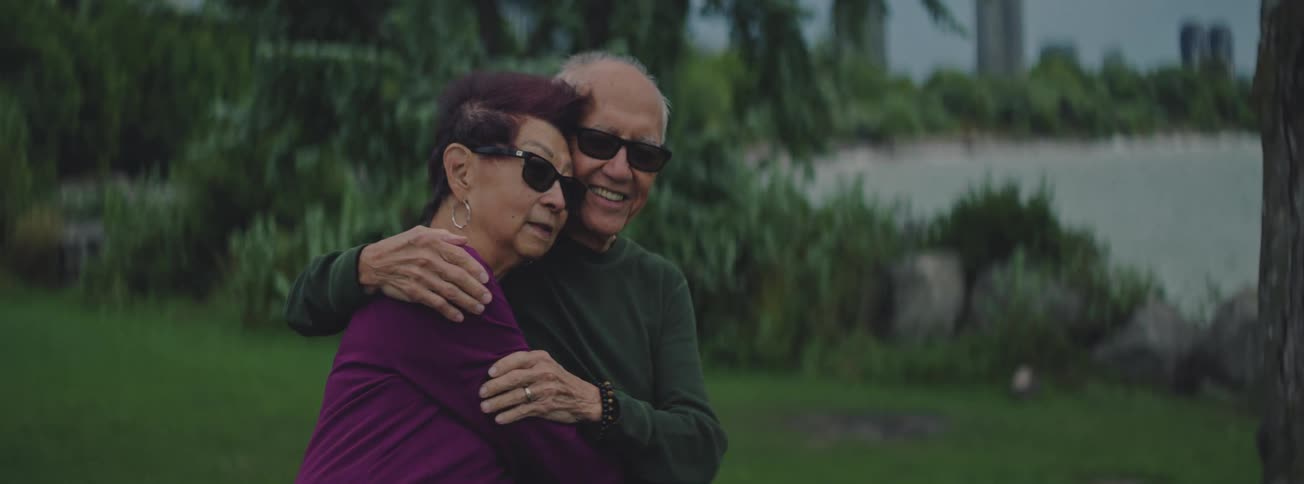 An elderly couple wearing sunglasses shares a loving embrace in a peaceful green park