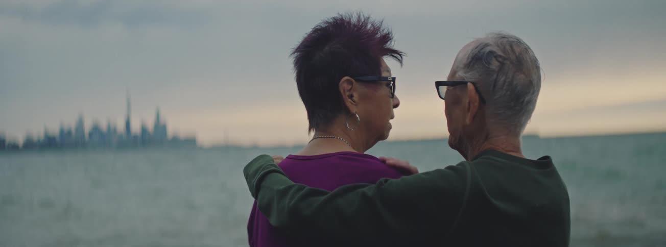 A loving elderly couple embraces while enjoying a quiet, contemplative moment together looking out over the water towards a distant city skyline