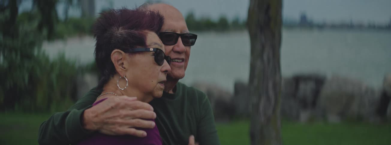 An intimate slow motion shot reveals an elderly couple sharing a loving embrace while sitting in a park by the water